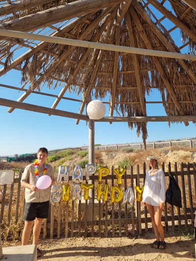 In Palmachim, Israel, two people pose for a photograph under a thatched-roof shelter, likely on a beach or near one. A man, wearing a Hawaiian shirt, lei, and dark shorts, stands on the left, holding a pink balloon. To his right, a woman in a white dress and flower crown stands. Behind them, a wooden fence displays "HAPPY BIRTHDAY" balloons, a mix of silver and gold. The environment is dry and sandy with sparse vegetation, indicating a coastal or arid climate. A clear blue sky dominates the background.