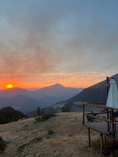 The image depicts a mountainous landscape at sunset. The sun, a bright orange orb, is partially obscured by a mountain range on the left side of the frame, casting a warm glow across the sky and clouds. The sky transitions from a bright orange near the horizon to a pale blue at the top. In the foreground, a wooden deck with a railing overlooks the valley. On the deck, there are two chairs and a table, and a white umbrella is folded and propped up. The ground around the deck is covered in dry grass and scattered rocks, with some evergreen trees visible on the slopes. The distant mountains are layered, with some appearing hazy in the fading light. The scene suggests a remote, natural setting, possibly a viewpoint or a resting spot in a mountain area near San Juan de Plan/San Chuan de Plan, Spain, given the provided context. There are no people visible in the image.