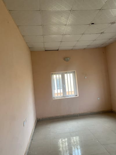 The image shows an interior view of an empty room in Bulamare, Nigeria. The room has light pink walls and a tiled floor in a neutral grey color. A white-framed window with vertical bars is visible on one wall, and it appears to be a double-pane window. On the same wall as the window, there are two electrical outlets. Another electrical outlet is visible on the opposite wall. The ceiling is made of square tiles, some of which appear to be damaged or discolored. A light fixture is attached to the ceiling near the window. The room appears to be recently painted or under renovation, as there are some drips of white paint along the base of the walls. There are no people or furniture visible in the room. The lighting suggests it is daytime, with natural light entering from the window.