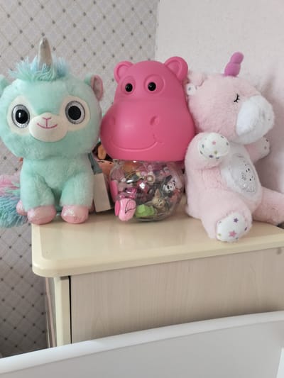 Three stuffed toys are arranged on a shelf. On the left is a light blue unicorn with a shimmering horn and large, sparkly eyes. To its right is a large, pink plastic hippo toy with round ears and black eyes. In front of the hippo is a clear plastic jar filled with small figurines, and behind it are more small stuffed toys, one of which appears to be brown. On the far right is a light pink unicorn with a purple horn, decorated with small colorful stars on its paws and body. The toys are on a light-colored wooden shelf with a smooth, rounded edge. Behind the toys is a wall with a subtle diamond-patterned wallpaper. The scene appears to be indoors, likely in a child's room or play area, given the presence of the stuffed animals and figurines. The lighting is bright, suggesting it is daytime, and there are no indicators of weather conditions. There is no visible text in the image.