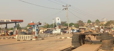 A roadside scene shows a gas station to the left with "DAMU OIL" on the canopy. In the center is a utility pole with power lines. Various shops and structures line the right side of the road. In the foreground, a makeshift structure is constructed from stacked tires and wood. People are seen walking and gathering near the shops and gas station. The scene takes place under a hazy, overcast sky.