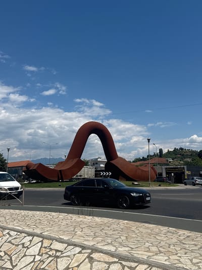 In the city of Shkoder, Albania, a massive rust-colored metal sculpture, resembling a giant horseshoe or clamp, dominates the scene. The sculpture is situated on a grassy roundabout, with a road encircling it. A black Audi sedan is captured mid-motion, driving on the road in front of the sculpture. To the left of the sculpture, a white van is partially visible, also on the road. Beyond the sculpture, a truck and other vehicles are parked or driving. In the background, buildings, hills, and a bright blue sky with scattered clouds complete the urban and natural landscape. The foreground features a textured stone sidewalk.