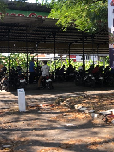 The image depicts a parking area with motorcycles parked under a covered structure. Several people are visible, some standing and others seated at tables. The environment appears to be outdoors, with trees and foliage in the background, suggesting a warm climate. The ground is paved and covered with fallen leaves. A white pillar is located in the foreground. In the distance, buildings and banners are partially visible. The scene suggests a public gathering or waiting area, possibly associated with a restaurant or market given the presence of seating and the covered parking. The context of Buduran, Indonesia, implies a tropical setting.