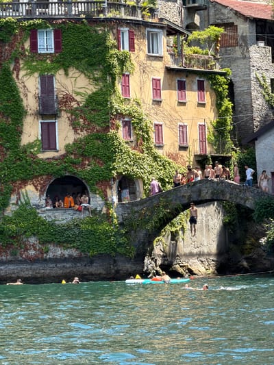 In the city of Nesso, Italy, an arched stone bridge spans a body of water. On the bridge, a group of people are gathered, some standing, some sitting. One individual is captured mid-air, having jumped from the bridge into the water below. The buildings on the left are covered in lush green ivy, with red-shuttered windows adding a splash of color. The water is a vibrant green, with several people swimming and a few inflatable rafts visible on the surface. The scene depicts a lively summer day with people enjoying recreational activities by the water.