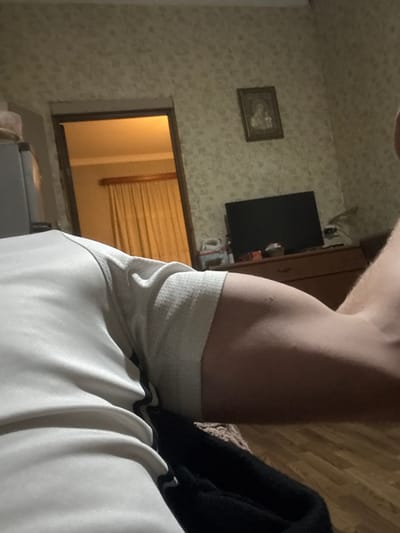 The scene is an interior residential room located in Durgena, Georgia. The image is captured from a low, first-person perspective. In the immediate foreground, a human arm, wearing a white short-sleeved garment with a black stripe detail, extends from the lower left towards the right. The arm displays visible bicep development and a small dark mole on its outer surface. A black fabric item is partially visible beneath the arm on the lower right. The room features patterned wallpaper on the rear wall. A wooden door frame on the left opens to an adjacent room, where a light-colored curtain covers a window, illuminated by an internal light source. A framed icon depicting religious figures is mounted on the wallpapered wall. Below it, a dark flat-screen television is positioned on a wooden dresser. Various small, indistinct objects are on the dresser to the left of the television. The floor is constructed of wood or laminate.
