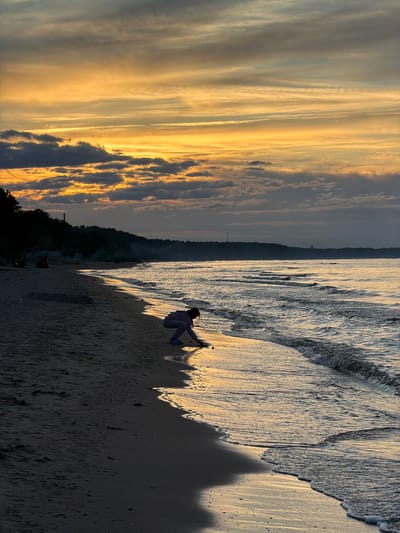 The image captures a serene beach scene at sunset in Plieņciems, Latvia. The sky is filled with dramatic clouds painted in hues of orange, yellow, and gray, indicative of the golden hour. The calm sea gently laps at the sandy shore, reflecting the warm light of the setting sun. In the foreground, a person is crouched on the wet sand near the water's edge, silhouetted against the bright reflection of the sky. They appear to be engaged in an activity, possibly drawing or playing in the sand. The silhouette of a tree-covered coastline stretches across the middle ground, with a faint structure visible on the horizon, suggesting a distant tower or lighthouse. The overall mood is peaceful and contemplative, amplified by the soft lighting and the vastness of the natural landscape.