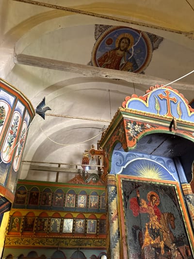Captured from a low-angle perspective within the church of Dolno Uino, Bulgaria, the image reveals vibrant and intricately painted religious iconography. A circular mural on the wall depicts a figure, likely a saint or religious figure, surrounded by a patterned border. Dominating the foreground are elaborately decorated elements of the church's interior, including a wooden structure adorned with floral patterns and religious symbols. Above, a section of the ceiling showcases an icon of a mounted figure, possibly Saint George, against a backdrop of radiant light. The overall composition emphasizes the ornate and devotional art within the sacred space.