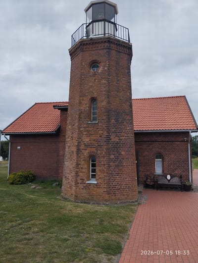 The image shows the Ventė Cape Lighthouse in Ventė, Lithuania. The lighthouse is a brick tower with a glass lantern room at the top. It is surrounded by a red-tiled roof building, also made of brick. The lighthouse has several windows and a balcony. In front of the building, there is a red brick pathway and a green lawn. A wooden bench is placed near the entrance of the building. The sky is overcast, and there are some trees visible in the background. A date and time stamp "2026-07-05 18:33" is visible in the lower right corner of the image. There are no people visible in the image.