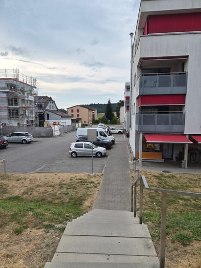 The image shows a street scene in Belfaux, Switzerland, on an overcast day. In the foreground, a concrete staircase with a metal railing leads down. To the right of the staircase is a patch of dry, brown grass interspersed with green. A paved walkway runs alongside the grass.

In the middle ground, there is a parking lot with several cars parked. A silver hatchback is parked on the right, and a darker car is parked behind it. A white van is also visible. A building with red awnings and balconies is situated on the right side of the image. The building has a modern design with white and grey facade.

Further back, there is a construction site with scaffolding surrounding a building. Several other residential buildings and trees are visible in the background. A stop sign is present near the construction site. The sky is cloudy.