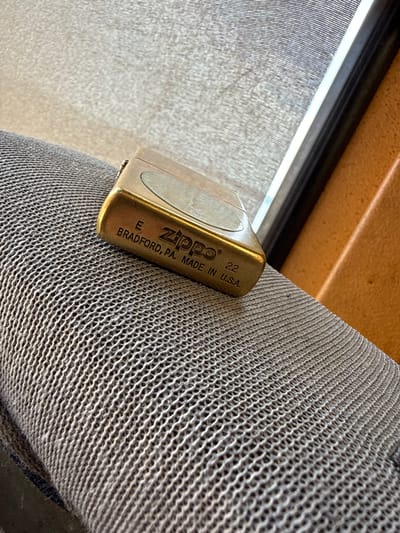 A Zippo lighter rests on a textured, grey surface, possibly a fabric or mesh material. The lighter is made of brass and has some text engraved on its surface. The text reads "ZIPPO 22 BRADFORD, PA. MADE IN U.S.A." and also "E". The lighting suggests it is daytime, with light filtering from an unseen window or opening. The background is blurred, showing a tan colored surface, and a dark, thin line that could be part of a frame or edge. The overall impression is one of a still life or object study, with the Zippo lighter as the central subject. The context of Mikołajki, Poland, suggests this might be a personal item within a home or outdoor setting in that location. There are no people or specific events depicted.