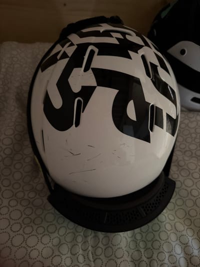 The indoor scene, reportedly situated in Bormio, Italy, features a white ski helmet prominently displayed. The helmet exhibits a bold, black geometric pattern covering its surface, possibly depicting abstract shapes or stylized letters. Multiple dark scratches and scuff marks are visible on the white shell, particularly across the upper left and central portions. The helmet features several elongated vertical ventilation slots. Its lower rim consists of a black, textured or vented band. The helmet rests on a light-colored fabric surface adorned with a repeating pattern of concentric circles. Partially visible to the upper right and behind the main subject is a second helmet, predominantly dark in color with white or light accents. The background beyond the fabric appears to be a light brown or beige solid surface, likely a wall or shelf.
