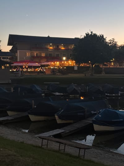The image depicts a twilight scene at the harbor in Prien am Chiemsee, Germany. In the foreground, several boats are moored, covered with dark blue tarps, resting on a pebbled shore. A wooden bench is visible in the lower right corner, placed on grass.
In the middle ground, a waterfront promenade is lined with tables and chairs, many covered by large, pink umbrellas. The lights from the buildings and lamps create a warm glow. A few people are visible walking or sitting, and a motorcycle is parked.
The background features a multi-story building with balconies, appearing to be a hotel or restaurant, with lights illuminating its facade and the surrounding trees. The sky is a soft gradient of blue and orange, characteristic of dusk, with a single bright star or planet visible in the upper left. The water in the harbor is calm, reflecting the lights.