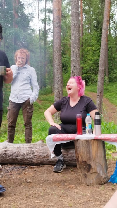 The image depicts a casual outdoor gathering in a forest setting, likely in Orekhovo, Russia, given the user prompt. In the foreground, a woman with vibrant pink hair is seated on a rustic wooden stump, laughing heartily. She is wearing a black t-shirt and black pants, with grey socks and black and white sneakers. On the stump in front of her are three cylindrical containers: a red thermos, a clear plastic bottle of water, and a silver thermos. A red and white patterned cloth is spread out on the stump, covered partially by a white plastic sheet. To the left of the woman, a person with wavy red hair is standing, holding a metal mug to their mouth, and wearing a light-colored button-down shirt and brown cargo pants. Smoke is visible behind them, suggesting a campfire or cooking activity nearby. Further to the left, another person is partially visible, wearing a black t-shirt and a black hat, holding a metal mug. The background consists of tall pine trees and lush green foliage, with a dirt path visible in the distance. The ground is covered with dry leaves and dirt.