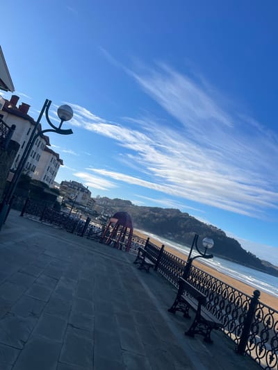 The image, captured with a distinct diagonal tilt, depicts a sunlit coastal promenade in Zarautz, Spain. In the foreground and midground, a wide pathway constructed of dark grey rectangular stone slabs extends from the lower left. This promenade is bordered on the right by a dark, decorative metal fence featuring repeating circular and oval patterns. Three dark-colored, traditional park benches with ornate cast-iron supports are spaced along this fence, facing the beach. Two dark-colored streetlights, each with a spherical lamp and curved support arms, are also visible along the promenade.

To the left of the promenade, multi-story buildings with light-colored facades and reddish-brown tiled roofs are visible, some featuring balconies with dark railings. On the beach side, a red, dome-shaped or arched structure stands in the midground, behind the fence. Beyond the promenade and fence lies a wide, light brown sandy beach meeting the blue-grey ocean, which exhibits visible white surf lines. The background is dominated by a large, tree-covered hill or mountain range rising from the coastline, with additional buildings nestled on its lower slopes. The sky above is a bright, clear blue, traversed by elongated, wispy white cirrus clouds. No distinct individuals are discernible in the scene.