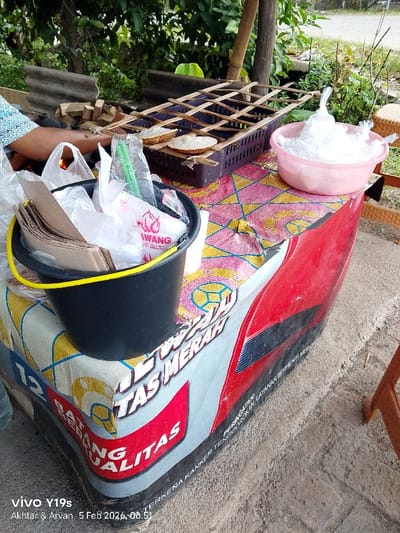 A vendor's food cart features a black bucket filled with brown paper bags, plastic bags, and green straws. Beside the bucket sits a metal grill holding small, round baked goods. A pink bowl filled with ice sits near the grill. The cart itself is decorated with the text "AWANG" and "BATANG KUALITAS TAS MERAH". The phrase "TERKENA KANKER TENGGOROKAN, LAYANAN BERHENTI MERD" is also visible on the cart. The background shows foliage and a partially visible structure.