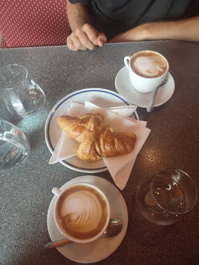 The image depicts a table scene in a cafe in Rovereto, Italy. A speckled dark grey table surface occupies the majority of the frame. Two individuals are partially visible at the top edge; one wears a red garment with white polka dots, the other a plain black short-sleeved shirt, with their forearms and hands resting on the table.
Arranged on the table are several items:
- Two white ceramic cups with matching saucers and metal spoons. Both cups contain cappuccino, distinguished by latte art: one exhibits a simple swirl, the other a more intricate feather-like pattern.
- A white ceramic plate with a blue double-line rim, upon which two golden-brown croissants rest on white paper napkins.
- Two clear glass tumblers, one on the far left partially filled with water, and one on the far right which is empty.
- A portion of a clear glass carafe or pitcher, also containing water, is visible on the left side of the table.
The scene suggests a breakfast or coffee break for two individuals.