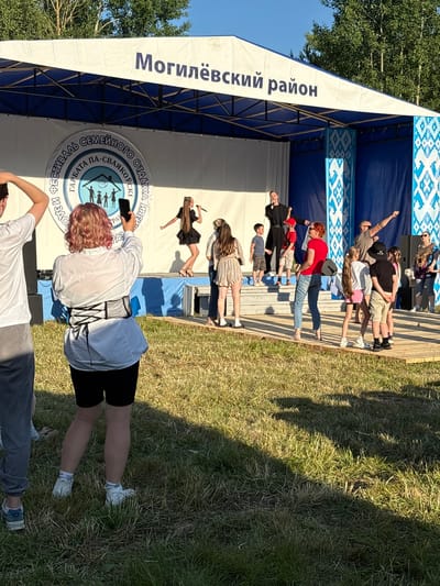 An outdoor stage event is underway in the city of Вяйнянскі сельскі Савет, Belarus. The stage backdrop features banners with Cyrillic text, including "Могилёвский район" (Mogilev Region) and a circular logo with the text "ФЕСТИВАЛЬ СЕМЕЙНОГО ОТДЫХА" (Family Recreation Festival). Several performers are on stage, including a woman singing into a microphone and a man in a black outfit also holding a microphone. A group of children and adults are watching the performance from the stage area and the audience. In the foreground, a person with pink hair is holding up a smartphone to record the event. The scene is set on a grassy area, with trees visible in the background. The lighting suggests it is daytime, with sunlight casting shadows on the ground.