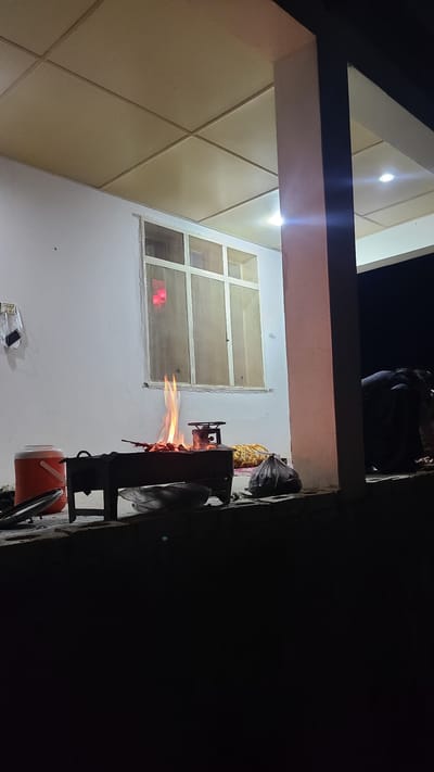 The image depicts a nighttime scene in what appears to be a residential area. A person is visible sitting on a structure above. Below, a grill with a visible flame is actively cooking food, with bags and other items nearby. A window is visible in the background. The description includes the location: Ziarat, Pakistan.