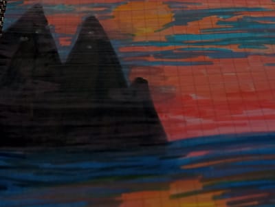 A colored drawing on squared paper depicts a mountainous landscape at sunset. Two dark, silhouetted mountains dominate the left side of the image. The sky behind them is a gradient of warm colors, transitioning from orange and yellow near the horizon to blues and reds, suggesting a sunset. The sun itself appears as a bright yellow orb partially obscured by stylized clouds. The foreground features a body of water, also rendered in shades of blue and reflecting the colors of the sky. The drawing appears to be created with markers or colored pencils, with visible strokes and grid lines from the paper showing through the coloring. The location in the city of Dymer, Ukraine is not discernible from the image.