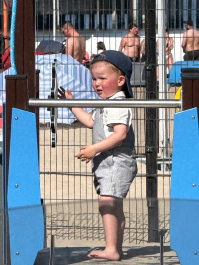 The image, captured in Blankenberge, Belgium, shows a young child lying horizontally on an outdoor play structure under bright sunlight. The child, likely a boy, wears a navy blue baseball cap, a white t-shirt, and grey and white striped shorts. His eyes are closed or looking downwards, and his cheeks appear flushed, possibly from sun exposure or exertion. He is barefoot, with his legs and feet extended to the right side of the frame.
A silver pole is positioned vertically near the child's head, and angular blue structural elements are visible above and below him, indicating components of a playground or recreational facility. A wire mesh fence or railing with a grid pattern is a prominent feature, creating layers and partially obscuring the background.
The ground surface beneath the structure appears light-colored, possibly concrete, with strong, defined shadows suggesting a sunny day, likely during midday or afternoon. In the background, several adults are visible, out of focus and partially obscured by the fence. One adult in a light blue and white striped shirt is seen behind the child, and another bare-chested person is in the upper part of the frame. The overall setting is an active outdoor recreational area. No text is visible in the image.