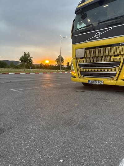A yellow Volvo semi-truck is parked on an asphalt lot. The truck is facing the viewer and is angled to the right. The sun is setting behind the truck, casting a warm glow on the scene. In the background, there are trees and hills. The license plate on the truck reads "CB 0273 CP". The scene is likely in Chuchuligovo, Bulgaria, as indicated.