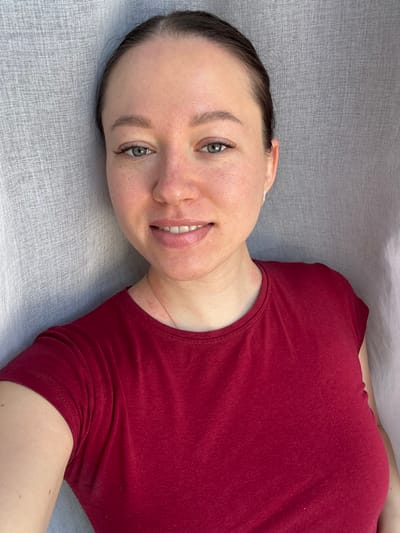 This image depicts a single adult female in an indoor setting within the city of Shul'gino, Russia. The subject, estimated to be between 25 and 35 years old, has fair skin with scattered freckles visible on her forehead, nose, and cheeks. Her dark hair is pulled back, revealing a clear hairline. She possesses greenish-blue eyes with enhanced eyelashes and well-defined eyebrows. Her lips have a glossy appearance, and she displays a closed-mouth smile, revealing her upper teeth. Small, silver-toned stud earrings are present in both earlobes. Erythema, consistent with skin irritation or a rash, is visible on her lower neck and upper chest area. She is wearing a solid dark red, short-sleeved, scoop-neck t-shirt. A thin, gold-toned chain necklace is partially visible around her neck. The subject is positioned directly facing the viewer, looking into the camera lens, indicative of a posed photograph or selfie, with her left upper arm partially visible in the lower-left corner of the frame. The background consists of a uniformly lit, light-colored (likely off-white or light grey) textured fabric, consistent with a curtain or drapery. No other objects or details are discernible in the immediate environment.