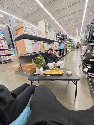 The image appears to be taken inside a retail store, possibly a large department store like Walmart, located in the city of National City, United States of America. The store is well-lit with overhead fluorescent lighting, illuminating rows of shelving stocked with various products.
A person wearing light-colored pants and a dark jacket is sitting in what seems to be a display setup. The setup includes a black folding table. On the table, there are various objects including a golden heart-shaped sculpture and a decorative glass bowl.
Shelves in the background are filled with items for sale, including storage cubes and desk components. There's also a display of children's books or toys on a lower shelf.
Another person is visible in the foreground, partially obscuring the view. They are wearing dark clothing, including leggings and a jacket. A blue object, possibly a bag or cushion, is also visible.
The overall scene suggests a candid moment within a commercial environment.