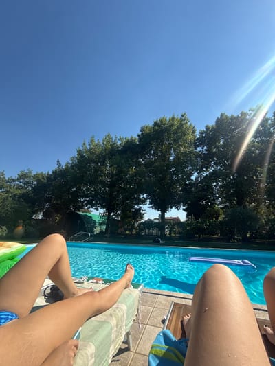 The image depicts a sunny outdoor scene at a swimming pool, likely in Reggio Emilia, Italy, given the context provided. Two individuals are seen from the waist down, lounging on chairs beside the pool. One person, closer to the viewer, has their legs extended and is holding a small electronic device. Their legs show stretch marks. The other person's legs are also visible, lying in a similar relaxed posture. The swimming pool is a vibrant blue, with ripples on the surface indicating movement. A purple inflatable mattress floats in the water. Behind the pool, lush green trees line the background, with a clear blue sky above. The sun is shining brightly, casting a lens flare into the upper right corner of the image. The environment suggests a leisurely day spent by the water.
