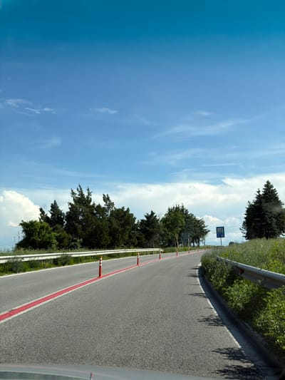 The image displays a bright, sunlit road scene, likely captured from inside a moving vehicle. The primary subject is a paved asphalt road, extending into the distance with a gentle curve. A prominent solid red line runs parallel to the right edge of the road, accompanied by several orange and white striped flexible bollards, suggesting a designated lane, possibly for cycling or specific traffic. To the right of the red line, a white solid line marks the road shoulder, beyond which a metal guardrail separates the road from abundant green bushes and grass.
On the left side of the road, and also further in the distance on the right, dark green coniferous trees are visible, contrasting with the vibrant green undergrowth. The sky above is a clear, bright blue with scattered white clouds, indicative of fair weather. Strong sunlight casts distinct shadows of the guardrail posts and nearby foliage onto the road surface on the right. In the distance, on the left side of the road, a blue rectangular road sign can be seen, though its white text and symbols are too blurry to decipher. The setting suggests a drive through a rural or semi-rural area near Brestovets, Bulgaria, on a sunny day. A reflection of the road on a vehicle's dashboard or window is visible in the bottom right corner.