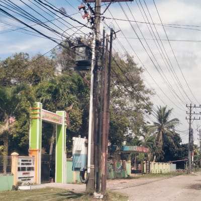 This daytime photograph, taken in Lapang Timu, Indonesia, depicts a roadside scene dominated by utility infrastructure and lush tropical vegetation. In the foreground, a prominent cluster of utility poles is heavily laden with a dense network of black electrical wires stretching across the frame against a clear, partly cloudy sky. A large grey transformer is conspicuously mounted on the central pole.
To the left, a colorful green and orange archway serves as an entrance, with an adjacent sign. Visible text on the archway and sign suggests it could be a school, possibly reading "SMA YAYASAN" or "SEKOLAH MENENGAH ATAS YAYASAN" in parts. Behind this structure, various trees including tall palm trees and other deciduous foliage contribute to the verdant landscape.
The unpaved road, made of dirt or gravel, extends towards the background where another smaller, faded blue and green archway and a light green and yellow fence are visible. Another wooden utility pole, also carrying wires, stands further down the road on the right. The bright light indicates mid-morning or afternoon, and the weather appears sunny and calm. The scene is static, devoid of people or active events, focusing instead on the interplay of natural growth and human-built infrastructure in a tropical setting.