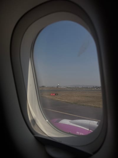 The image captures a view from inside an airplane, looking out through a porthole-style window. The dark, rounded frame of the window occupies the foreground, framing the scene outside.
Outside the window, a light blue, slightly hazy sky fills the upper left portion of the frame. Below this, the landscape unfolds, dominated by a dry, yellowish-brown field or scrubland. On the right side of the view, part of the airplane's engine cowling is prominently visible. It features a distinctive color scheme of purple and silver-white sections. On the purple section, the white text "NO STEP" is clearly visible, indicating a safety instruction.
Beneath the engine and extending into the middle distance, an asphalt surface, likely a taxiway or runway, can be seen. Further in the distance, a low-lying urban or industrial area with buildings is present, backed by hazy mountains or hills on the horizon. A white, spherical structure, possibly a radar dome or a similar facility, stands on the ground in the mid-distance. To its right, a rectangular red sign with white text is visible on the ground. The text on this sign reads "IL-29R A10," which appears to be an airport designation for a runway or taxiway.
The lighting suggests it is daytime, possibly mid-morning or afternoon, with a bright but somewhat diffused light due to the hazy sky. The overall impression is that the aircraft is on the ground at an airport, likely taxiing, waiting for takeoff, or having just landed.