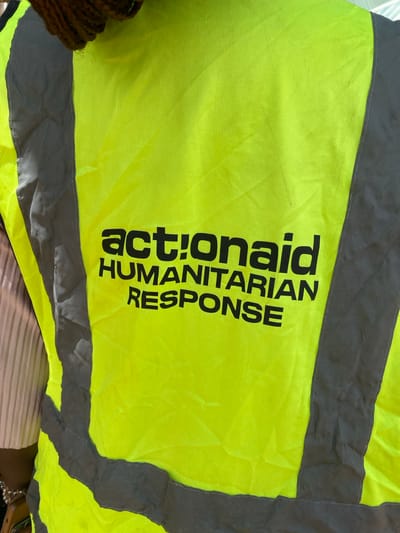 The image shows the back of a person wearing a bright yellow safety vest with reflective grey stripes. Black lettering on the vest reads "act!onaid HUMANITARIAN RESPONSE". The person's hair is visible at the top, woven in braids. Part of a pink garment is visible under the vest. The background is out of focus, suggesting an outdoor environment. No specific location within Rim, Nigeria is identifiable from the image.
