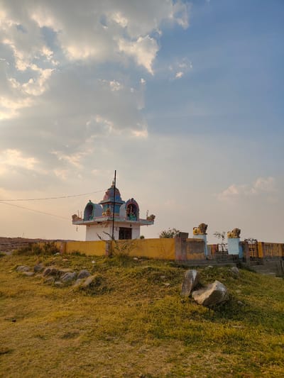 The image captures a small Hindu temple located in Kammavaripalli, India. The temple is a white, square structure with a colorfully decorated roof. The roof includes pink, blue, and green paint. There are two lion statues on either side of the roof. A yellow wall with some graffiti surrounds the temple. The temple sits on a grassy hill dotted with rocks. A wrought iron gate leads to the temple from a small set of stairs. The sky above is a pale blue with scattered white clouds. There are no people visible in the scene. Electrical lines can be seen cutting across the upper portion of the image.