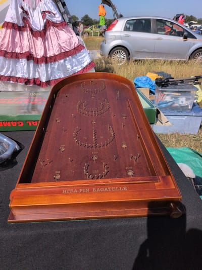 The image depicts a vintage wooden bagatelle game set on a black surface, likely a table or stall at an outdoor market. The game board is made of polished wood and features a series of brass pins and several circular holes with numerical scoring values printed around them, including 50, 60, 70, 90, 100, 125, and 150. The text "HIT-A-PIN BAGATELLE" is inscribed on the front of the wooden frame.
In the background, a large, ruffled pink and white dress is visible, suggesting a costume or theatrical prop. Behind the dress, a silver car is parked on a grassy area. Further in the distance, other vehicles and people can be seen, as well as trees and a bright blue sky, indicating an outdoor setting. A green box with red lettering that appears to say "JACQUES" and "AL GAM" is positioned to the left of the bagatelle game. Other items, including what appears to be a blue tarp, a clear plastic container with various objects, and a tripod, are also present in the background.
The overall scene suggests a fair, fete, or car boot sale in an open, possibly rural or suburban environment. While the exact location within Claverley, United Kingdom is not explicitly identifiable from the visual elements, the context of an outdoor market is strongly suggested.