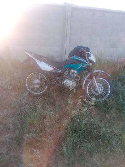 A dirt-bike is parked in a grassy, dry area near a concrete fence. The dirt-bike is teal and white with a black seat and has a backpack strapped to the handlebars. The bike has a "YAQİ" logo on the seat and "250" on the side panel. The sun is low in the sky, casting a bright glare on the left side of the image, suggesting it is either sunrise or sunset. The weather appears dry and warm, typical of a steppe environment. The scene is likely rural, given the natural vegetation and the simple fence. No people are visible, and the overall mood is peaceful and still.