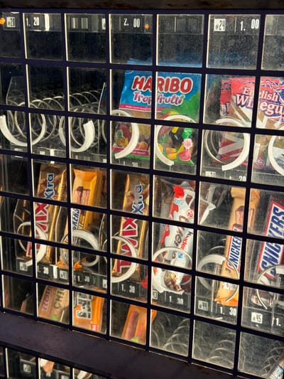 The image shows a section of a vending machine filled with various candy bars and bags of sweets, likely located in Witten, Germany, as indicated by the prices displayed in Euros. The machine has a grid of glass compartments, each containing a product and a corresponding price label.
In the top row, visible products include a bag of Haribo Trop Froot, and two bags of what appear to be English sweets. Prices shown are 1.20, 2.00, and 1.00.
The second row displays several chocolate bars. Brands identifiable include "Xtra," and "Kinder" (likely Kinder Chocolate). Prices for this row range from 1.30.
The third row also contains candy bars, including what appears to be Snickers bars. Prices are consistently 1.30 in this section.
The vending machine has a metallic outer frame and a dark blue background behind the products. The overall condition of the machine appears to be functional, though the glass displays show some dust and smudges. No people are present in the image. The lighting suggests it might be daytime, possibly with artificial light from within the machine.