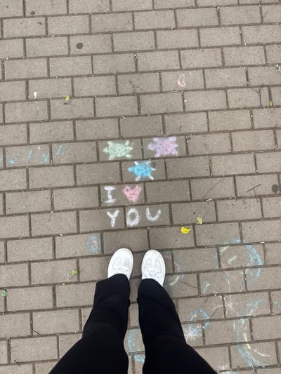 This image captures a close-up, overhead view of a paved sidewalk in Chastyye, Russia, marked with chalk drawings. The sidewalk is composed of rectangular gray bricks. In the center of the frame, chalk art spells out "I LOVE YOU" with an "I" in white, a heart symbol in pink, and "YOU" in white. Above this message, there are chalk drawings of what appear to be turtles in light green and light blue. In the foreground, the lower legs and feet of a person wearing black pants and white sneakers are visible, standing on the sidewalk. Scattered chalk markings in various colors, including pink, blue, and green, are present across the pavement, along with a few fallen leaves and twigs.