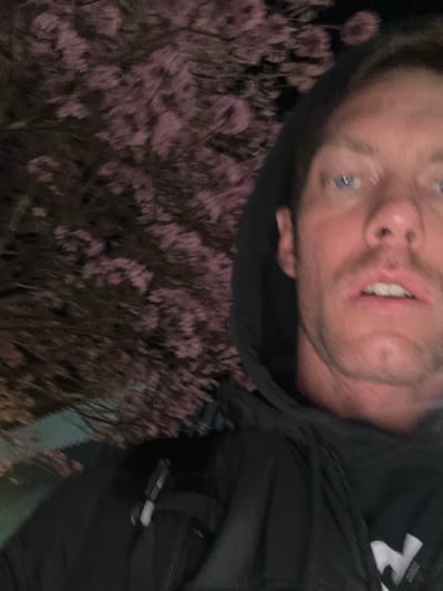 A male individual is depicted in a close-up, likely a selfie, in Long Beach, United States of America, during nighttime conditions. He possesses blue eyes, light brown hair, and facial stubble, looking directly forward with his mouth slightly ajar. He is wearing a dark hooded jacket, with the hood pulled over his head. A white object, possibly a pen or clip, is attached to the left side of his upper garment. A partial white graphic is visible on the lower right front of his jacket. The background is dominated by a tree in bloom, featuring numerous small pink blossoms, dimly lit against a dark sky. A faint greenish light source is visible in the lower left background, beneath the blossoms.