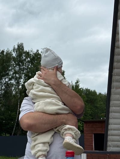 A man, wearing a white t-shirt, is holding a baby wrapped in a cream-colored fleece outfit and a white hat. The baby is facing away from the camera, looking towards the right. In the background, there are green trees under a cloudy sky. To the right, part of a wooden structure with horizontal siding and some decorative string lights is visible. A small, dark wooden shed with an open doorway is situated behind the man. The scene appears to be outdoors, possibly in a rural or suburban area.
