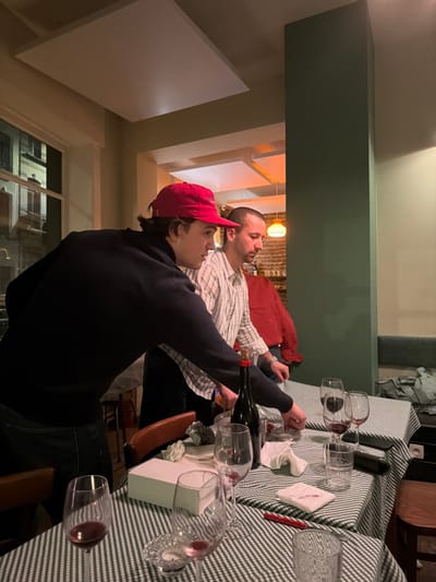 An interior scene captured within a commercial establishment in Ixelles - Elsene, Belgium, displays two adult males interacting with items on tables. The foreground features a male wearing a red baseball cap and a dark blue long-sleeved top, leaning forward with his right arm extended over a table. Behind and to his right, a second male, attired in a light-colored striped shirt, also leans over the table.

The tables are draped with green and white checkered tablecloths. Visible items include several wine glasses, most containing residual red wine; a dark-colored bottle with a cork stopper; a white rectangular box; crumpled white paper or napkins; a red pen; a clear drinking glass; and a small, clear glass object, possibly a candle holder.

The environment includes light-colored walls, a prominent dark green vertical structural element, and multiple white rectangular acoustic panels affixed to the ceiling. A large window on the left reveals reflections of exterior buildings, indicative of an urban setting. In the background, an orange spherical pendant light illuminates a section of wall constructed from exposed brick. The overall lighting is artificial and subdued.