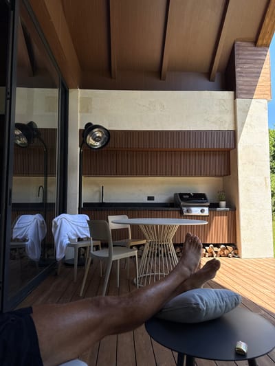 The image captures an outdoor living space, likely a patio or balcony, in Bakovići, Montenegro. The viewpoint is from a reclining position, showing a person's legs and feet resting on a light gray pillow placed on a dark gray round table. The person is wearing black shorts.
The background reveals a modern outdoor kitchen area with dark wood cabinetry and slatted paneling. A grill is positioned on the countertop. Next to the grill, a small potted plant is visible. Below the kitchen counter, a neat stack of firewood is stored.
To the left, a large glass door or window reflects the scene, including two chairs with white towels draped over them and a modern standing heater. The floor of the patio is constructed from wooden planks. The ceiling above is also wooden, with exposed beams. The wall behind the kitchen area is textured and light-colored. The overall aesthetic is contemporary and designed for outdoor relaxation and entertainment. The sunlight suggests it is daytime, casting shadows across the wooden floor and the wall.