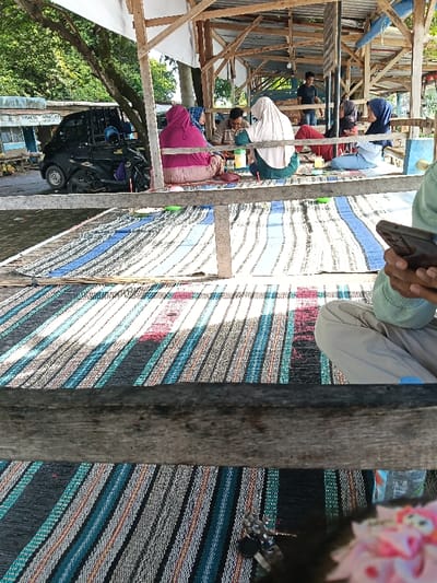 The scene depicts an outdoor or semi-outdoor communal area in Gembong, Indonesia, structured with visible wooden posts and beams supporting a partial roof of white fabric and corrugated metal sheets. The floor is covered with multiple striped mats of varying colors, including blue, white, black, red, and green.
In the immediate foreground, the lower body of an individual is visible, seated on a multi-colored striped mat. This person wears light-colored cargo pants with a blue accent on the right thigh and a light green long-sleeved top. Their hands are occupied holding a dark-colored smartphone. A set of silver keys and a blurred pink object are on the mat near their feet.
In the midground, five individuals are seated directly on various patterned mats. From left to right: an individual in a bright pink hijab and matching top, an individual in a blue patterned hijab, a male in a striped polo shirt, an individual in a light cream hijab with a dark green top (facing partially away), and an individual in a dark blue hijab with a light blue top. Small plastic containers (green, yellow, red) are positioned on the mats in front of them, suggesting a meal or social gathering. A clear plastic water bottle is also visible on the mat.
In the background, a black mini-truck is parked, with a black scooter positioned in front of it. Beyond these vehicles, a street is visible, and piles of coconuts are present on the ground near buildings, one featuring a blue facade. Further within the wooden structure, another male individual wearing a dark t-shirt stands. The environment is illuminated by natural daylight.