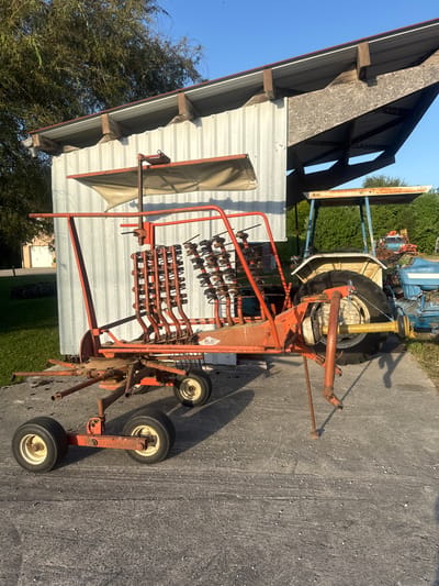 An outdoor, eye-level, wide shot shows a red, six-tined hay rake parked on a concrete surface. The rake is positioned in front of a metallic, corrugated building with a sloped roof. A portion of a white tractor with a blue roof is visible behind the rake. Several other farming vehicles, including a blue tractor and a red tractor with a crane, are in the background. Green trees and a clear blue sky form the backdrop of the scene. The image does not contain any people or evidence of recent activity.