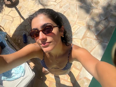 A woman is lying on her back outdoors, possibly at a swimming pool or a beach, given the presence of the water and the tiling. She is wearing a striped bikini and sunglasses, and her skin is wet, suggesting she has recently been in the water or is sunbathing after swimming. Her arms are spread wide, and her head is tilted back, with her face partially illuminated by sunlight. The background shows a mosaic-tiled surface and a portion of a lounge chair with a blue towel. Shadows from overhead foliage are cast on the ground. The overall impression is one of relaxation and enjoyment on a sunny day. The time of day appears to be daytime, with bright sunlight casting strong shadows. There is no visible text in the image.