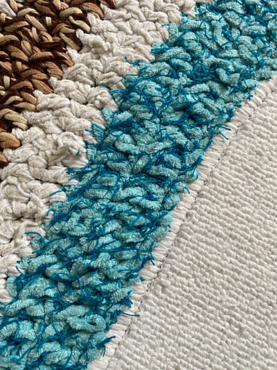 The image shows a close-up of a textured rug with a diagonal stripe pattern. The rug features sections of thick, woven brown and beige yarn, followed by plush white yarn, then a vibrant turquoise yarn with loose blue threads, and finally a more tightly woven white section. The lighting suggests an indoor setting, likely in a home or studio. The specific location in Osek, Czechia, is not discernible from the image.