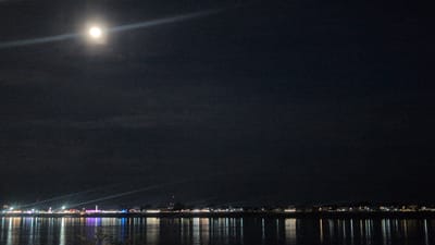 This is a nighttime photograph of the Mekong River in Vientiane Capital, Laos. The full moon is visible in the upper left portion of the sky, casting a bright light that creates a visible beam through the clouds. The opposite bank of the river is illuminated by a string of lights, likely from buildings and businesses. The lights are reflected in the dark water, creating a shimmering effect. There are no people or specific objects clearly identifiable in the image. The overall scene depicts a calm and atmospheric night along the river.