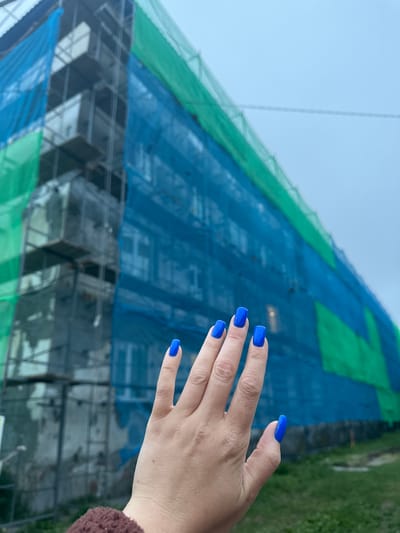 A person's hand with bright blue fingernails is held up in the foreground. Behind the hand, a multi-story building undergoing renovation is covered in scaffolding and green and blue netting. The sky is overcast. The scene is likely a residential building in Vilyuchinsk, Russia, given the context of the request, though the specific location is not visually verifiable within the image itself.