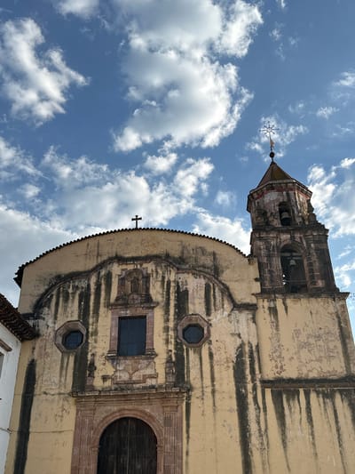 An upward-angled medium shot depicts the historic facade of a weathered, light-yellow church in Pátzcuaro, Mexico, against a backdrop of a partly cloudy, bright blue sky. The church's facade features an aged texture with noticeable wear, as evidenced by dark vertical streaks indicating water damage. Architectural details include an arched doorway on the lower right, rectangular and octagonal windows, and decorative carvings. A smaller cross is mounted above one of the windows. The top of the church culminates in a bell tower with openings to reveal visible bells inside, surmounted by a metal cross. White cumulus clouds fill the sky.