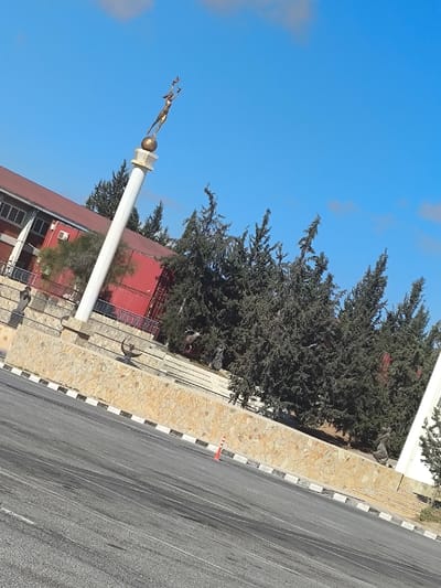 The image captures a static outdoor scene in Kızılbaş, Northern Cyprus / Cyprus, under a clear blue sky with sparse white clouds. The primary subject is a multi-tiered monument complex observed from a low angle, slightly tilted to the right.
In the foreground, a section of dark asphalt road exhibits minor surface variations, with a solitary orange traffic cone positioned near its right edge. This road is bordered by a black and white checkered curb. Directly behind the curb, a robust retaining wall constructed from irregular, light-colored stone blocks forms a raised platform.
Upon this platform and within its immediate vicinity, multiple elements are present:
A prominent tall, white cylindrical column with a golden capital rises from the stone base. It is topped by a golden-colored statue depicting an athletic female figure balanced on a sphere, extending her arms upwards, with a small bird (possibly a dove) in her left hand, suggesting an act of release or reception.
Several dark green coniferous trees are densely planted behind the stone wall, partially obscuring the background structures. Among these trees and on the stone terraces, several other sculptures are visible: two abstract human-like figures on the left, a deer with antlers situated amidst the trees, and another human-like figure partially visible on the far right.
In the background, behind the monument and trees, a multi-story building with a red roof and light-colored walls is visible on the left, featuring dark-framed windows and an external air conditioning unit. To its right, a large red structure, possibly a shipping container or a building with red corrugated siding, is also partially visible. No individuals are present, and no overt actions or interactions are depicted within the frame.