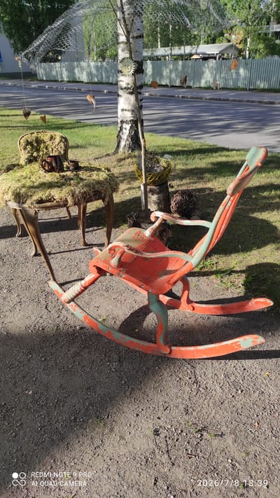 This image depicts an outdoor scene in the city of Iisalmi, Finland. In the foreground, a weathered, orange and teal rocking chair sits on a gravel surface. The chair has a distressed paint finish, showing signs of age and wear. To the left of the rocking chair is a small, ornate table covered in moss, with a brown ceramic mug and cup placed on top. Behind the table, a birch tree stands prominently, adorned with a decorative wire mesh canopy and several hanging ornaments, including heart shapes and a sun face. The background shows a residential street with a light-colored wooden fence and trees. The overall setting suggests a whimsical, outdoor art installation or decorative display.